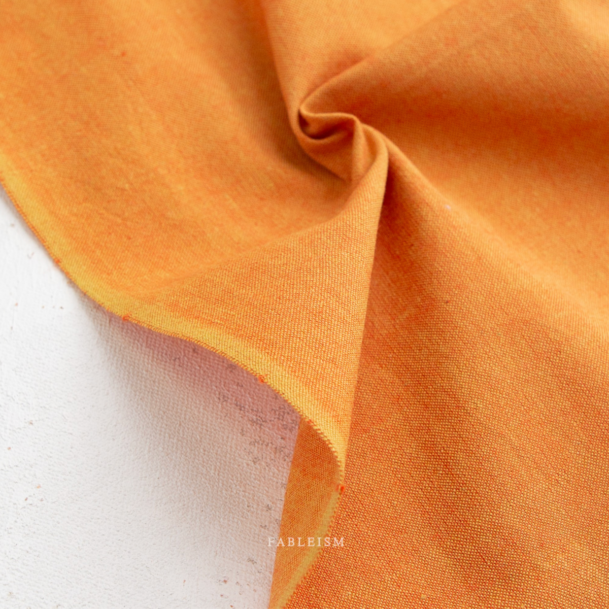A close-up of draped mustard yellow fabric with a visible woven texture, folded over to show the material and color against a light neutral background.