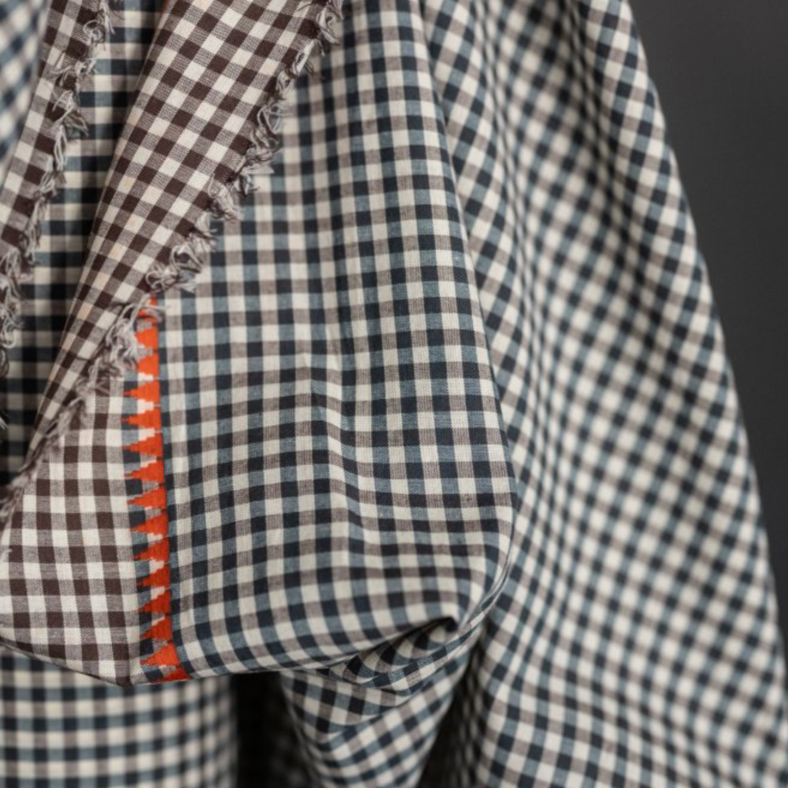 Close-up of a checkered fabric garment or textile with a brown and white gingham pattern, frayed edges, and a visible orange embroidered  accent.