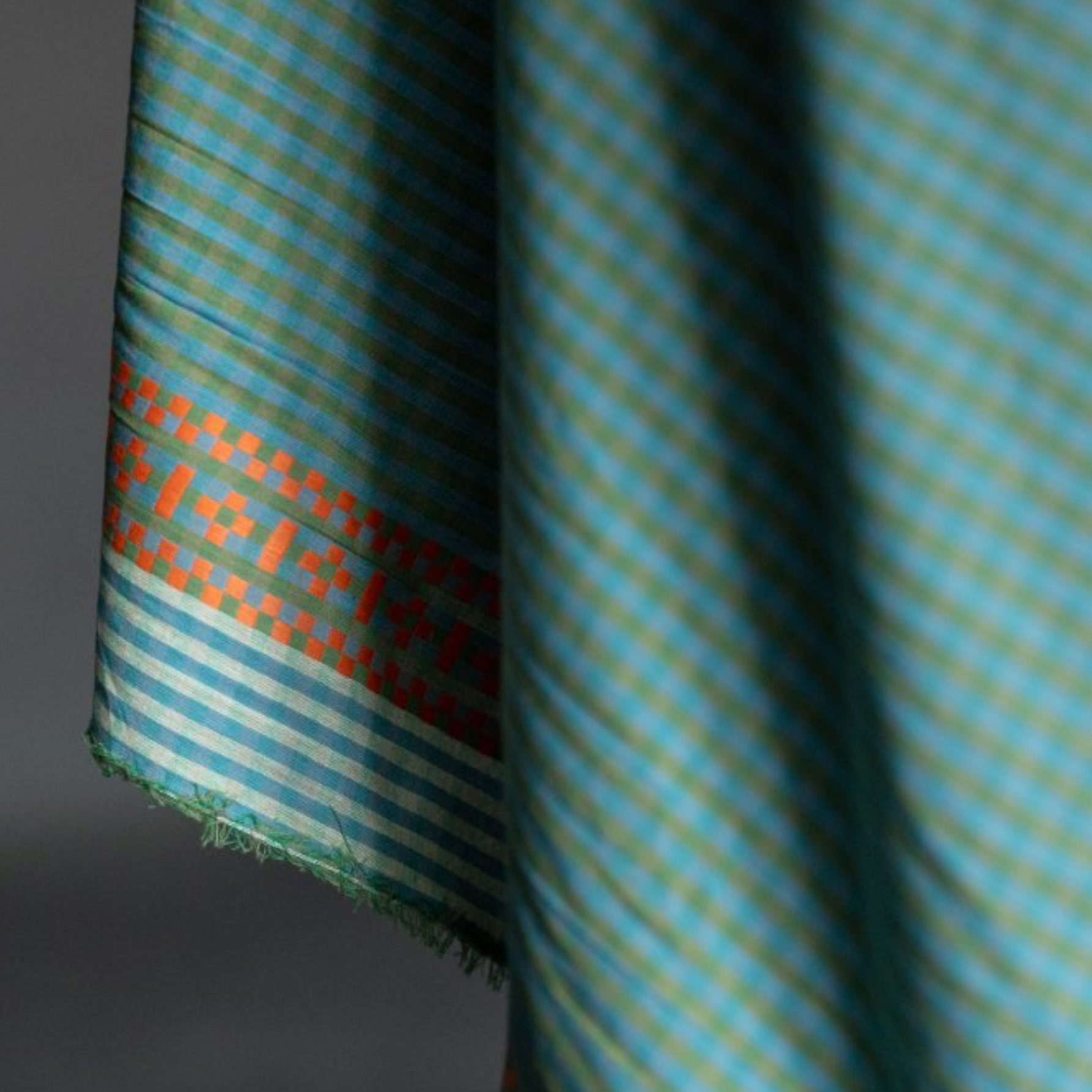 A close-up of afolded fabric featuring a teal and light blue checkered pattern with a decorative border of orange and green motifs.